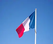 A vertical tricolour with blue, white and red stripes flies from a white flagpole before a clear blue sky.