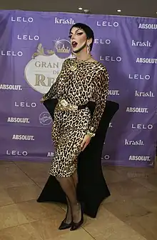 Venedita Von Däsh wearing a leopard print gown in front of a backdrop advertising Gran Hotel de las Reinas