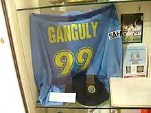 A blue coloured T-shirt displayed at a store window. The T-shirt has the words "Ganguly" and the number 99 below it, both in yellow colour. Beside the T-shirt, a picture and an open book is visible.