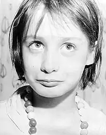 A clearly defined black-and-white close-up photograph of Genie from the chest up against an indeterminate background. Her eyes are focused slightly above and to the right of the camera, and she has an expressionless, vacant, innocent look on her face.