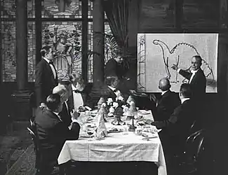 A black and white film still.  A group of men sit around a dining table in the center.  To the right, a man stands by and gestures at a large drawing of a dinosaur.