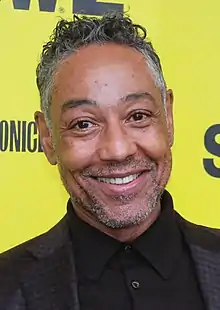 A smiling man with great and black hair and facial ahir, wearing a black buttoned shirt and patterned black blazer, looks directly into the camera.