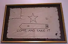 A white flag, with a five-pointed-star over the outline of a small cannon, with the words Come and Take It below.  The left corner of the flag has been torn off and sewn back on.