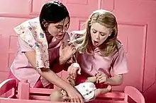 Two young women face forwards, standing over a pink wooden cradle. Both are wearing pink outfits and the background is a pink room. The woman on the left has chin-length dark hair and holds in her right hand a plastic lifelike baby doll. With her left hand she pushes the other woman, who has long, light brown hair and also leans over the doll.