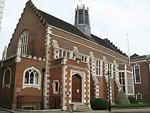 A large brick rectangular building, with a darker tiled sloping roof; the corners and buttresses of the building have larger white stone blocks as edging, instead of bricks. On the end of the longer side, a porch (approximately half the height of the main building), with four steps leading up to an arch and a large wooden door; above the door, a coat of arms of a golden griffin on a black shield; the wall of the longer side of the building has five brick buttresses, four small high windows and one large oriel window