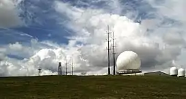 Photo shows a large white golfball-style radar dome, with two smaller radar domes and a number of other antenna; all set against a dramatic cloudy sky.