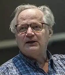 Color photograph of older Caucasian man with grey hair wearing blue flannel shirt and glasses.