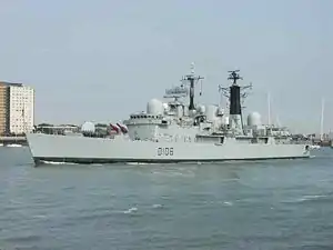 Grey warship with black towers and red missiles on its bow, city buildings are in the background.