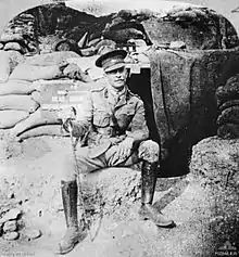 Soldier wearing Sam Browne belt and peaked cap, with a walking stick, sitting in front of a sandbagged doorway half covered by a tarpaulin. A sign says: "headquarters".