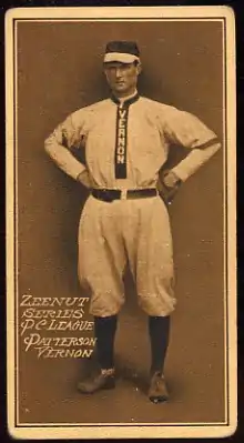 A baseball card of a man in a light colored baseball uniform with the word "Vernon" going down the center stands with his hands on his hips.