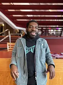 A photo Hanif Abdurraqib wearing a denim jacket over a black hoodie looking off camera and leaning against a counter.