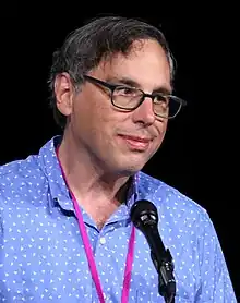 Harry McCracken sitting on a stage in a suit, gesturing with his hands while speaking