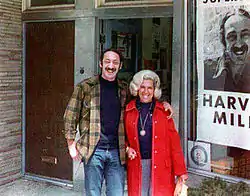 A color photograph of Harvey Milk and Audrey Milk standing in front of a store, with a campaign poster featuring a photograph of Harvey Milk in the background, partially out of frame