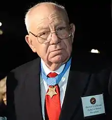 Head and shoulders of a gray-haired white man wearing glasses, a suit and tie, and a star-shaped medal hanging from a blue ribbon around his neck.