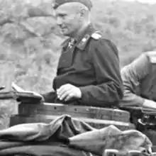 The head and shoulders of a man, shown from the side. He wears a black military uniform and cap, and an Iron Cross displayed at the front of his dark uniform collar.