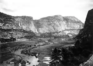 High cliffs bound a valley with meandering stream in it. One of the cliffs has a waterfall and another cliff is much larger than the others.