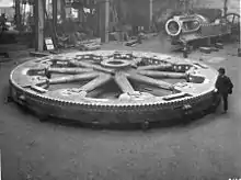 Flywheel for a large rolling mill engine 1900; the heavy rim is cast in four sections bolted together at the rim. Top right, the trunk guide and bedplate of the engine under manufacture, beyond the bedplate is the flywheel and connecting rod of a small horizontal steam engine.