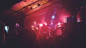 From a dark, crowded audience, the camera looks up at three performers standing before microphones, lit from behind with red light.