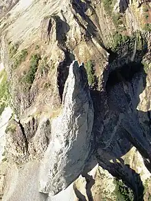 An aerial view of a vertical grey-coloured rock column rising above sparsely vegetated slopes.