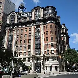 View of the Belleclaire Hotel from the intersection of Broadway and 77th Street. The facade is largely made of red brick, except at the base, where it is made of stone. The top of the hotel contains windows within a black mansard roof.