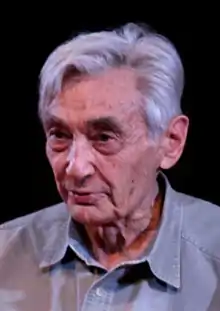 A color photograph of an older man in front of a black background, shot from his chest up