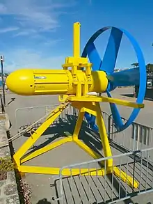 The Sabella D03 turbine. Painted bright yellow, a steel frame tripod supports the nacelle containing the generator. To the right, painted blue is the 6-bladed rotor, the tips of the blade connected by a metal band. The turbine is sat on a quayside with metal barriers around it.