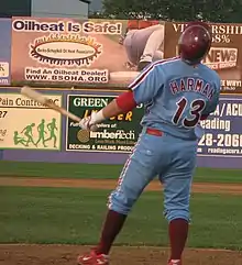 A man in a light-blue baseball uniform with maroon trim reading "Harman" and "13" on the back; he is holding a baseball bat in his left had and wearing a maroon baseball helmet atop his head