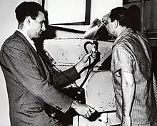 A photograph of showing two men standing over equipment on a table; one pours molten lead into the apparatus, while another holds an instrument connected to a pipe over it.