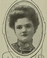 A portrait of a young white woman in an oval frame; she is wearing a high-collared blouse or dress, and her hair is in a bouffant updo. Her eyes appear to have been outlined with ink.