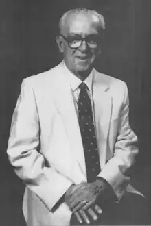 An elderly Jack Devine, seated with hand on lap, wearing a white suit jacket