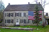 A large stone house with a large American flag draped across its upper right story behind an evergreen tree. In front of it is a blue and gold historical marker.