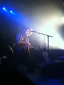 Ukulele player James Hill sitting on stage with a prepared ukulele (modified with a chopstick) on his lap.