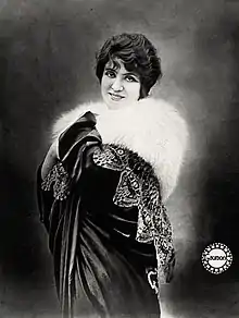 A smiling white woman with dark hair and eyes, wearing a white fur wrap over a dark draped velvet cloak