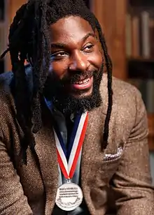 A man dressed in brown looks to the right side for the camera. On his neck is a ribbon with red, white, and blue stripes, with a silver medal attached to it.