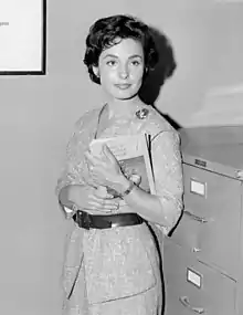 A woman holds a clipboard while standing next to a filing cabinet.