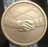 A bronze coin depicting a handshake with an axe and pipe crossed above it. "Peace and friendship" are written in text.