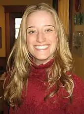 A young caucasian woman with blond hair and a red turtleneck sweater smiles at the camera