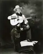 Studio picture of Jimmie Rodgers smiling and wearing cowboy attire: he dons a white Stetson hat, a short scarf on his neck, a two-tone western shirt, leather wrist cuffs, leather chaps, a revolver on the right side of his hips, cowboy boots, and spurs. A guitar hangs on his right shoulder from a strap as he holds it horizontally with his right arm. His left arm rests on his raised left knee and he holds a cigarette with his left hand. While his left foot rests on a stool covered with rugs his right leg stands straight