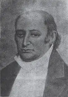 A man with dark, receding hair wearing a high-collared white shirt and dark jacket