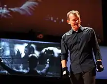 A man wearing a gray shirt, brow pants and black gloves stands in front of a screen, where scenes from Minority Report are being projected.