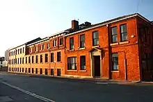 Large two and three storey building of red brick stretching into distance with modern vertical tube grain silos in distance having tarmac road to front and bright blue sky overhead