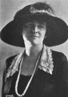 A white woman wearing a very large, wide-brimmed black hat, and a loose-fitting jacket with lace collar, and white beads or pearls