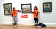 Two people wear orange "Just Stop Oil" t-shirts over black long-sleeve shirts. Each has a hand glued to the frame of a painting.