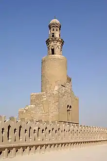 Photo of a minaret in four levels, with battlements in the foreground