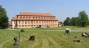 A photo at a slight horizontal angle of an old hunting lodge. It has three floors, pinkish to light orange walls, and a dark red roof. It is surrounded by a field and some gravel paths, with scultpures and a couple of cars standing on the grass at some places.