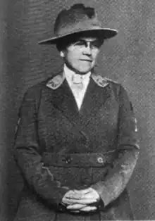 Middle-aged white woman, wearing glasses and a uniform-style jacket and hat, with hands folded in front of her body