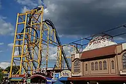 View of the Steel Curtain roller coaster from a nearby path. The roller coaster contains yellow supports and black track. A building is to the right.