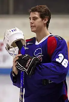 An ice hockey player standing, holding is ice hockey stick with his helmet rested on the top. He has short brown hair and is wearing a blue uniform.