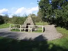 A pyramidal stone structure stands in a small clearing surrounded by small trees and bushes. The structure, enclosed by a fence, has an opening in the front.