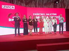 Nine persons visible chanting on a stage with "Lewica" branding in the back